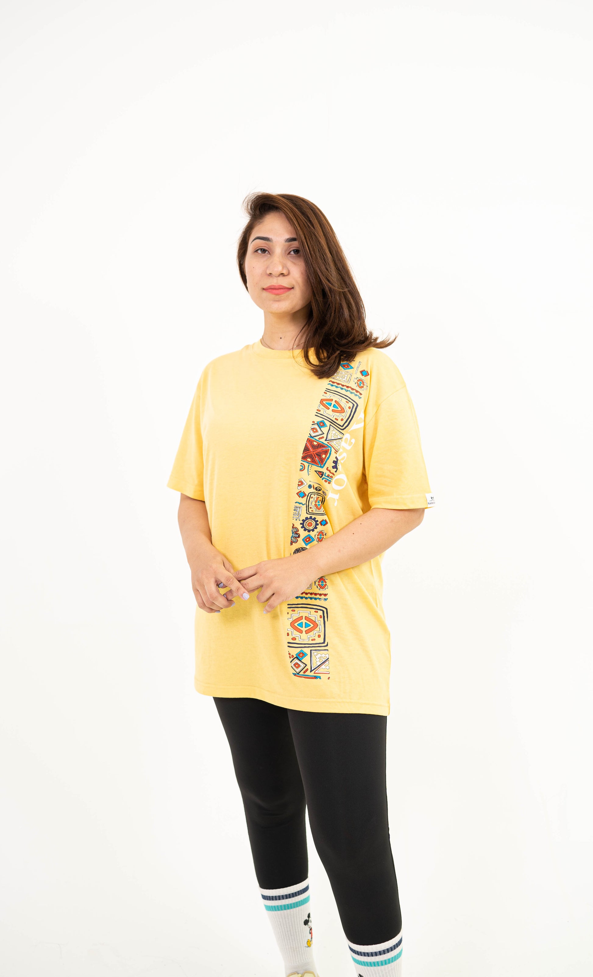 A woman modeling a yellow short-sleeve t-shirt with a colorful geometric pattern running down the front, paired with black leggings and white socks, against a plain white background.